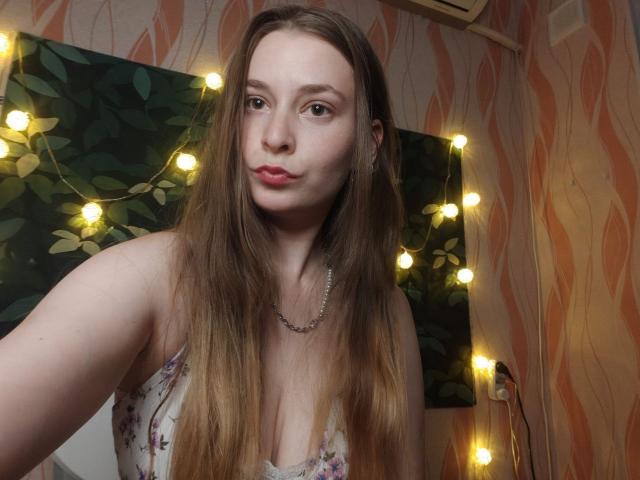 LunaBliss69 with long brown hair, pouty lips, and a necklace, wearing a floral dress, has small tits and a cute face, standing in a room with fairy lights.