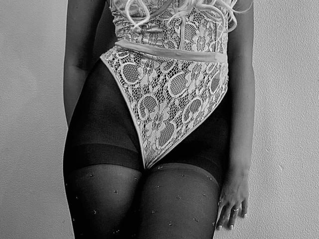 PrimeS wearing a lace bodysuit with a diamond pattern, covering their pussy, and dark, sheer, dotted tights, with blonde curly hair, standing against a plain wall.