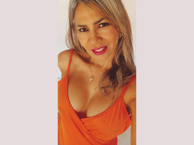 BrigiteWalsh has tanned skin, long blonde hair, and is wearing an orange dress with a low neckline, showing her boobs. She's smiling with red lipstick.