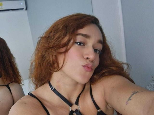 ValeryHilton with curly red hair, black harness, nose ring, and tattoo on right arm, pouting lips, wearing black strappy top, selfie in mirror.