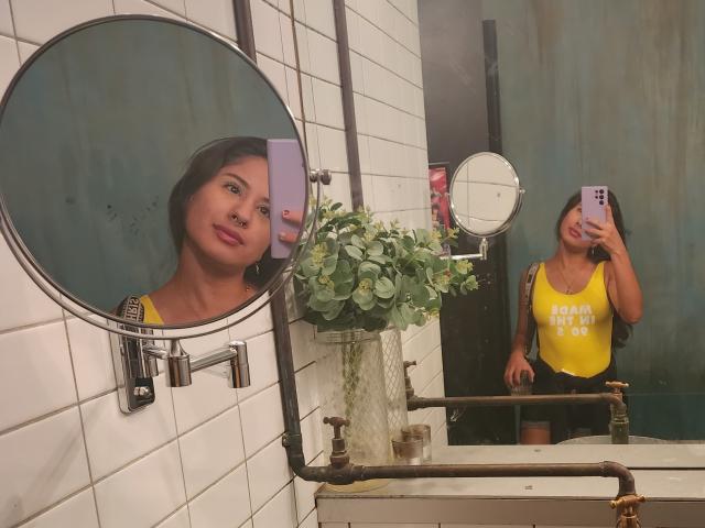 AuroraMonroe's reflection shows her smiling with a nose ring, wearing a yellow tank top and black shorts, holding a phone in a bathroom mirror selfie. Boobs are visible.