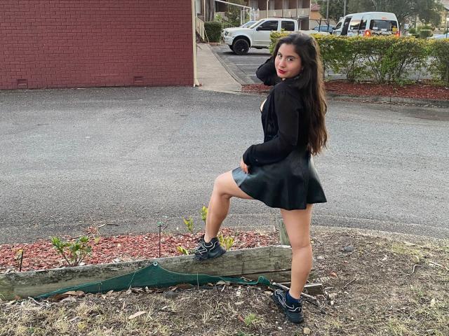 AuroraMonroe with long brown hair, black sheer top, green skirt, and blue sneakers, standing with one leg on a wooden plank, looking back with red lipstick.