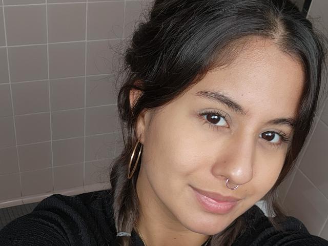 AuroraMonroe has medium brown skin, dark brown eyes, a small nose ring, and wears gold hoop earrings. She's smiling slightly with pink lips and dark hair in a messy bun.
