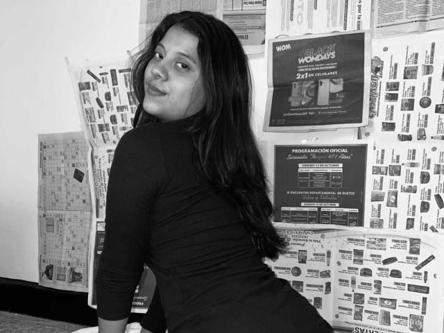 CoralineHale with long dark hair, black long-sleeve top, and ass facing camera, looking back with a slight smile, surrounded by newspaper-covered wall. Black and white photo.