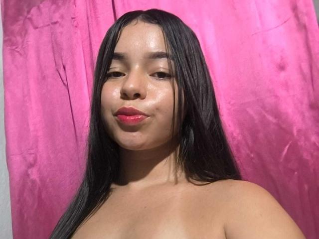 LissyCarter has long black hair, shiny skin, and red lipstick, with small, visible tits, and is topless, standing against a pink curtain, giving a slight pout.