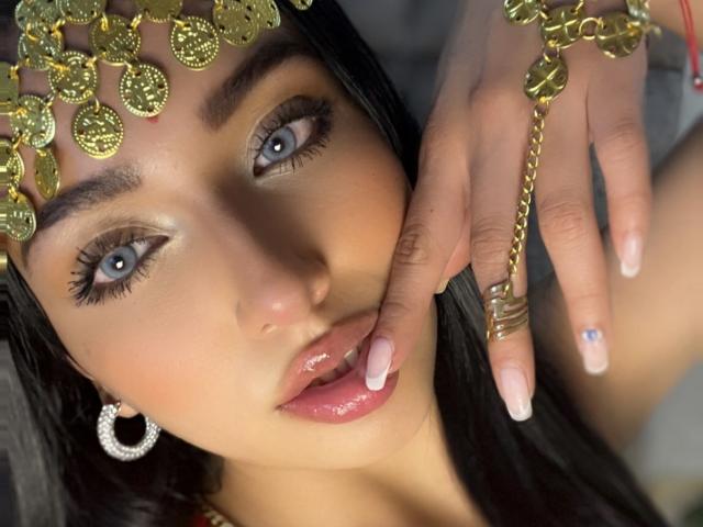 LillyTaylor's face, with blue eyes, glossy pink lips, and black hair, wearing a gold headpiece with circular ornaments, and a gold ring with a chain.