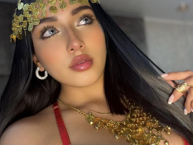 LillyTaylor with long black hair, blue eyes, and glossy pink lips, wearing a gold headpiece, red dress, and gold necklace, looking up with her hand near her face.