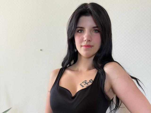 MarriAnni has long black hair, green eyes, and a small tattoo above her tits. She's wearing a black V-neck top, with pink lipstick and hoop earrings.