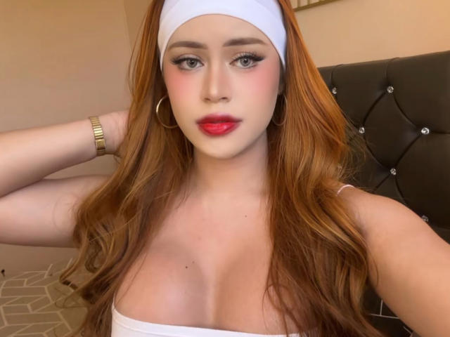 HeavenCummer with red hair, white headband, hoop earrings, and gold bracelet, wearing a white top, showing her tits, with red lipstick, and blue eyes in a selfie.