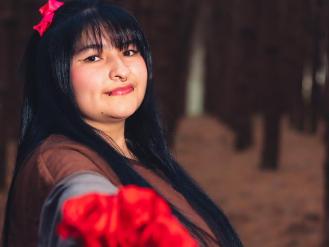 RadiantBebe with long black hair, pink bow, red gloves, and brown shirt, smiling slightly, reaching forward, cute, Asian, soft lighting, forest background, subtle nose ring, confident pose.
