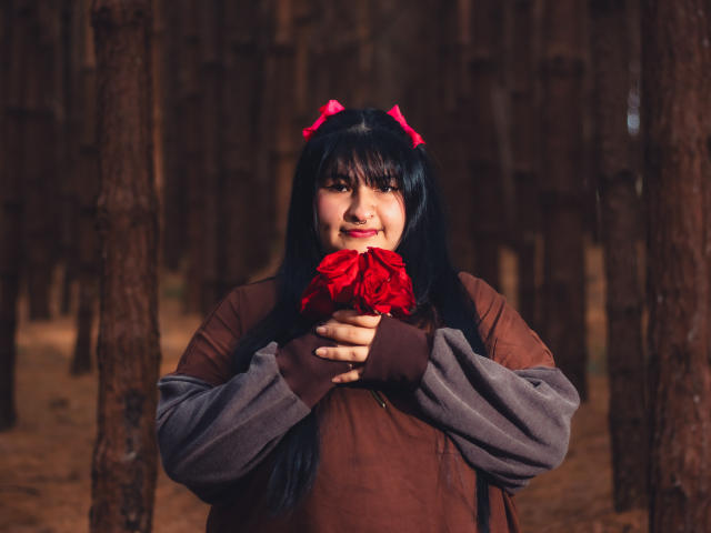 RadiantBebe with black hair, pink bow, brown jacket, and gray sleeves, holding red roses, smiling, in a forest. Her hands are on the roses, showing off her tits.