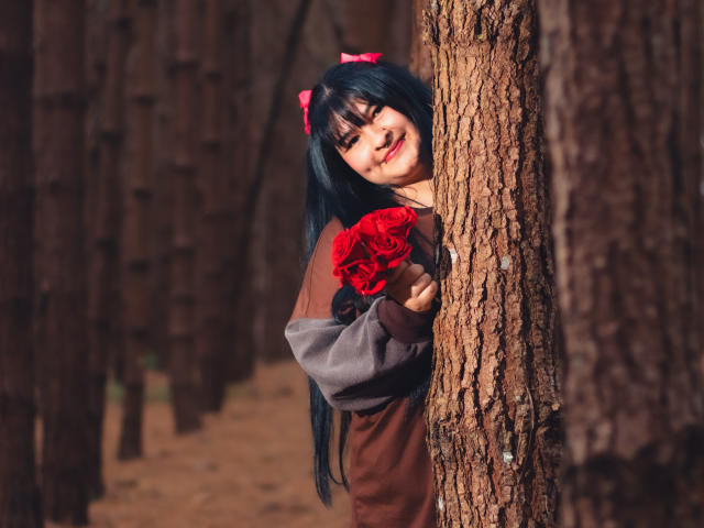 RadiantBebe with black hair, red bow, brown and gray sweater, holding red roses, peeks from a tree in a forest, smiling with red lipstick, long hair, cute.