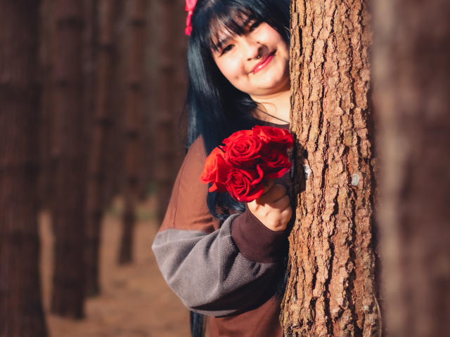 RadiantBebe with black hair, red bow, and brown-gray sweater, holding red roses, peeks from a tree in a forest, smiling softly.
