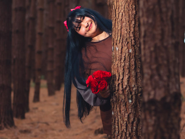 RadiantBebe with long blue hair, red bow, brown shirt, hiding behind a tree, holding red roses, smiling, forest background, thick tree trunks, cute, sexy, playful.