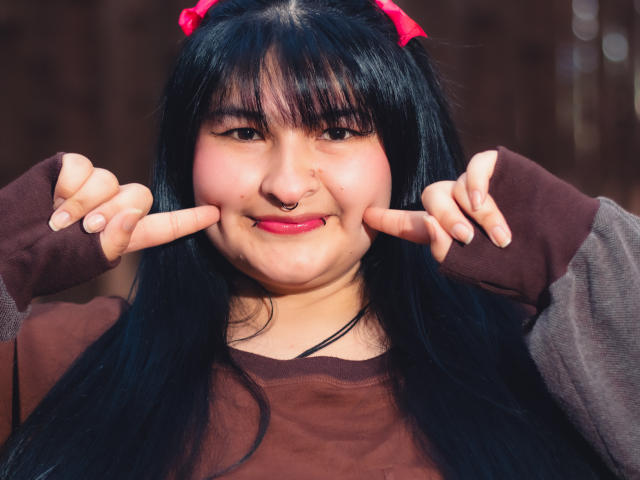 RadiantBebe with black hair, pink bow, nose ring, and brown shirt, making a double peace sign, smiling with pink lipstick, and showing a chubby face.