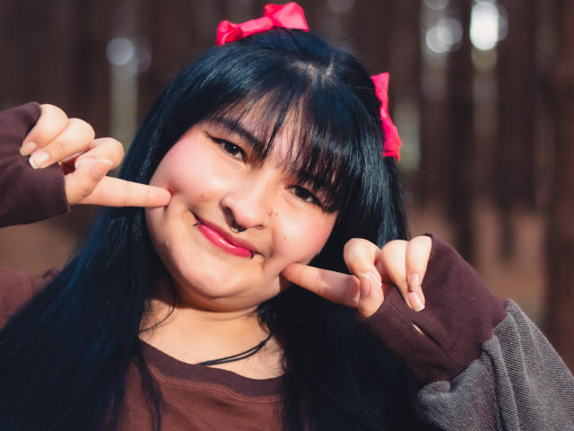 RadiantBebe, a chubby Asian girl with black hair, red bow, nose ring, and brown sweater, making double peace signs with a cute smile. Her pink lips are slightly parted.