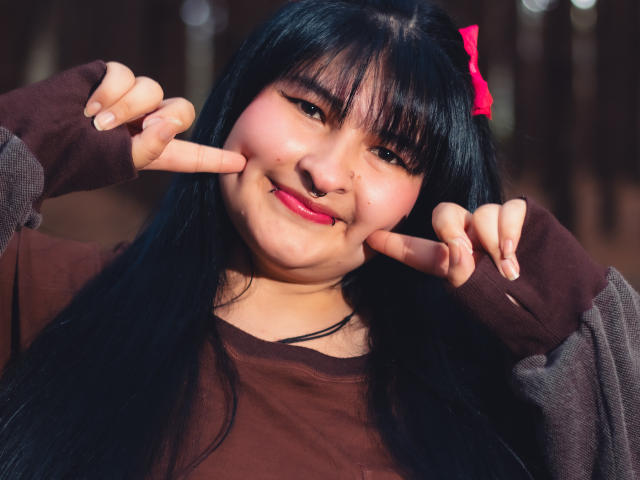 RadiantBebe with black hair, red bow, brown shirt, and finger guns, smiling with pink lipstick, chubby cheeks, and cute expression. Soft light on her face, slightly blurred background.
