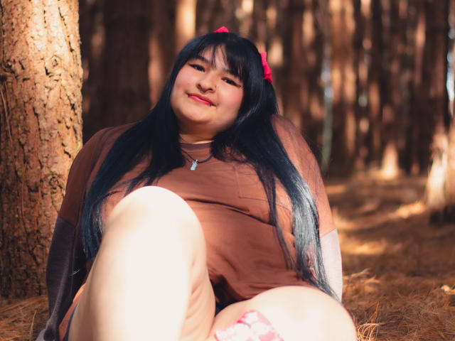 RadiantBebe, with long black hair and red bow, sitting against a tree, wearing a brown top, smiling, showing her ass, in a forest with sunlight filtering through.
