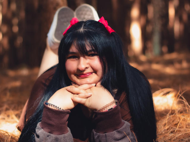 RadiantBebe with black pigtails, red bow, brown sweater, and hands under chin, smiling, lying on forest floor, legs up, sneakers visible, soft light, slightly chubby face.