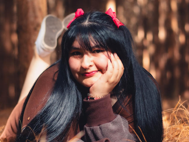 RadiantBebe with black hair, red bows, nose ring, and pink lipstick, laying in forest, brown jacket, hand on cheek, smiling, legs up, white sneakers, sunlit, cute.