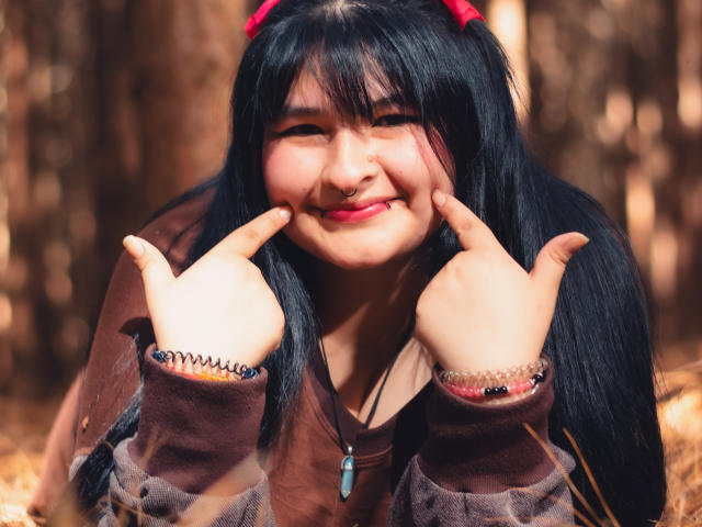 RadiantBebe with black pigtails, brown hoodie, and red hair ties, smiling and making "rock on" hand signs, has a cute nose ring and colorful bracelets.