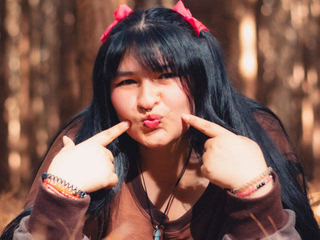 RadiantBebe with black hair and red bows, pouting her lips, pointing fingers at them, wearing a brown sweater, in a forest, sunlight filtering through trees.