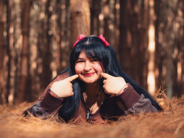 RadiantBebe with black hair, pink bows, and brown jacket, lying in pine needles, smiling with fingers pointing at her chubby cheeks, forest background, cute and playful.