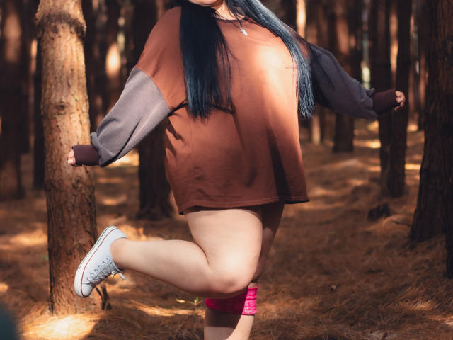 RadiantBebe with blue hair, brown and gray oversized shirt, pink shorts, and white sneakers, standing on one leg in a forest, arms out, touching trees.