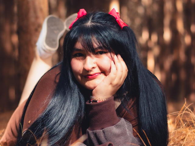 RadiantBebe with black hair, red bows, brown jacket, nose ring, smiling, lying in forest, hand on cheek, white sneakers, legs up, sunny, cute, fluffy, sexy.