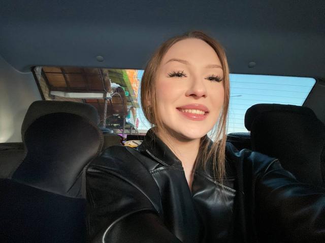 GabiCrystal with long brown hair, black leather jacket, and big eyelashes, smiling in a car, showing off glossy pink lips. Her tits are slightly visible under the jacket.