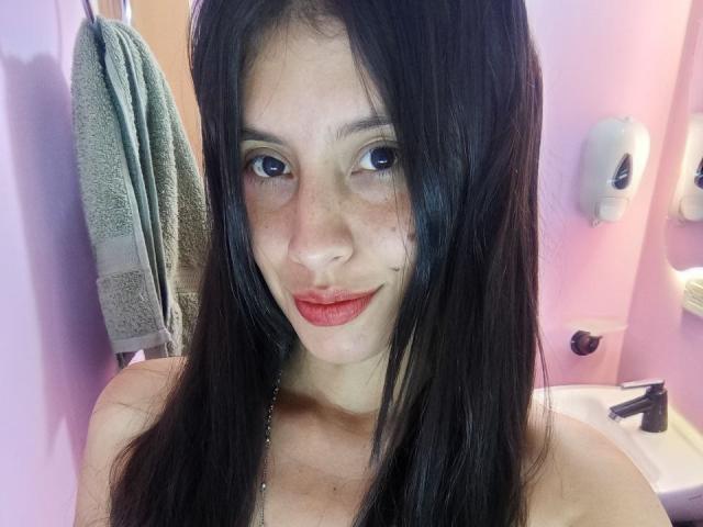 MadissonFontaine with long black hair, freckled face, and red lipstick, smiling at the camera, showing bare shoulders in a pink-lit bathroom. Her tits are not visible.