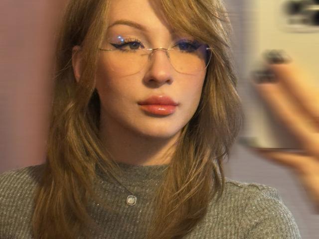 GabiCrystal with shiny glasses, pinkish lips, and wavy brown hair, wearing a grey ribbed sweater, showing off her pouty ass-kissing face in a soft, warm-lit photo.