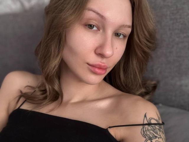GabiCrystal with wavy brown hair, light skin, and a small tattoo on her right shoulder, wearing a black strap top, puckering her pink lips slightly.