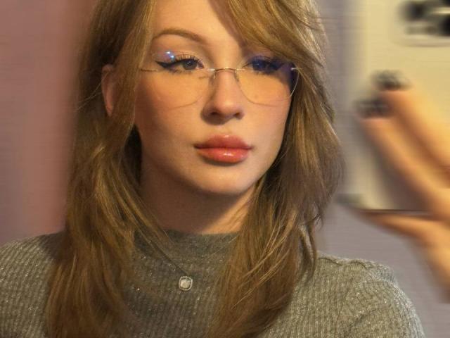 GabiCrystal has shiny pink lips, light brown wavy hair, and wears thin glasses with blue highlights. She's in a grey ribbed top, looking slightly off-camera.