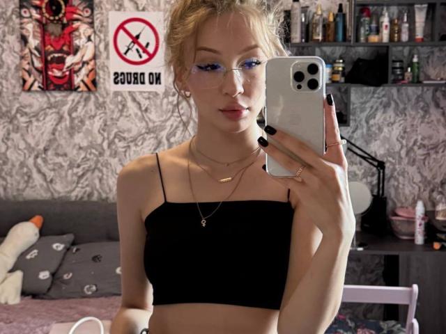 GabiCrystal with blue eyeliner, glasses, black tube top, blonde updo, holding iPhone, mirror selfie, black nail polish, necklace, bedroom with shelves, stuffed animal, and wall art.