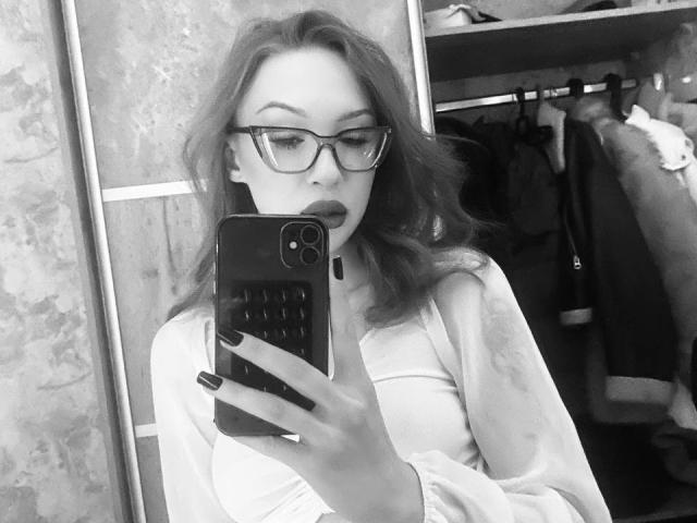 GabiCrystal with glasses, wavy hair, and black lipstick, holding a phone, looking down, wearing a light shirt, showing her tits in a black-and-white mirror selfie.