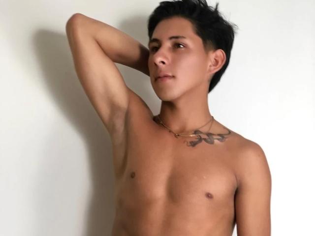 SoffiaVegga, shirtless, with short black hair, arm raised, showing a tattoo on the chest, wearing a necklace, brown eyes, looking to the side, against a plain white background.