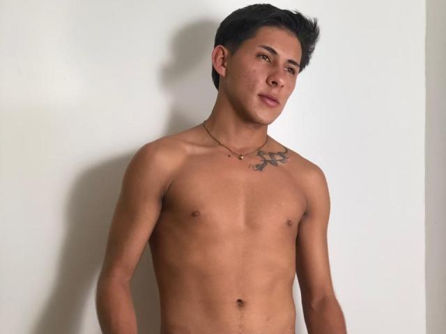 SoffiaVegga is a shirtless dude with tan skin, short black hair, a chest tattoo, and a necklace, standing against a white wall. He's looking to the side.