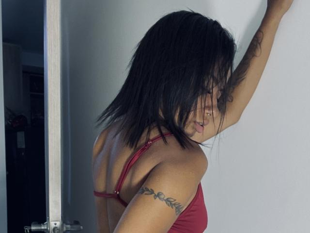 AhteneaSexy, con capelli neri a metà schiena, reggiseno rosso, tatuaggio sul braccio destro, piercing al naso e uno sguardo di lato, appoggiata a un muro bianco, che mostra il suo seno piccolo..