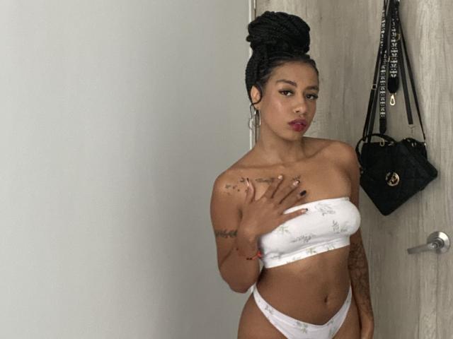 AhteneaSexy with dark skin, braided hair in a bun, wearing a white strapless top and white panties, hand on chest, black bag on door, tattoos on arm.