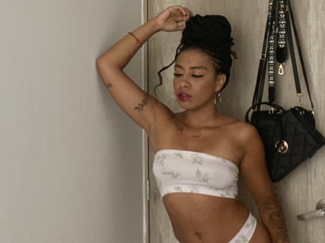 AhteneaSexy, avec sa peau foncée et ses cheveux tressés, ajuste son haut, portant un bandeau blanc sans bretelles et un bas assorti, un bracelet rouge, des tatouages, et un sac noir accroché à un mur en bois..