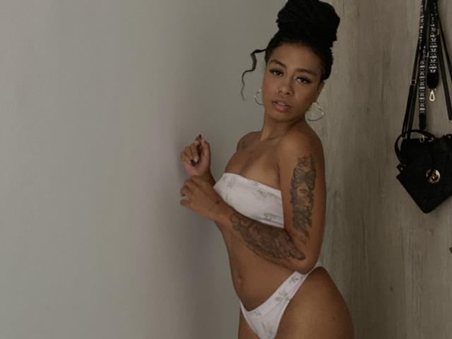 AhteneaSexy, con piel oscura, recogido negro, bikini blanco sin tirantes, brazo tatuado, de pie contra una pared, mirando a la cámara, bolso negro a la derecha, aros grandes, pose sexy..