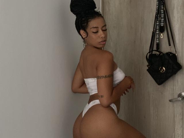 AhteneaSexy mit dunkler Haut und hochgesteckten Zöpfen, trägt weiße, trägerlose Unterwäsche mit String, wobei ihr Hintern und ihre kleinen Brüste zu sehen sind. Sie hat die Arme verschränkt und steht neben einer schwarzen Tasche..