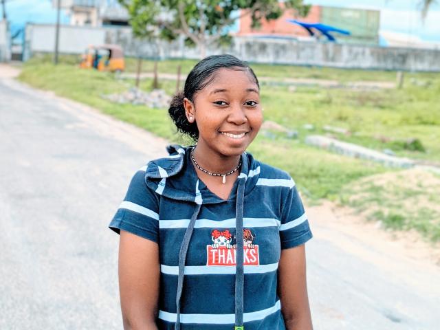 MariaThomson with dark skin, black hair in a bun, smiling, wearing a blue and white striped shirt, necklace, and hood, standing on a road. Her tits are small.