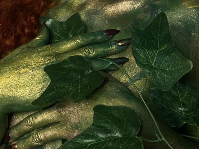Sahrataylorr's green-painted hands with black nails, covered in dark green leaves, resting on their golden-green painted face. Sahrataylorr has red curly hair, and the leaves are large.