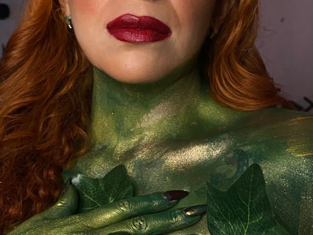 Sahrataylorr has green painted skin, shiny and textured, covering from neck to hand with green leaves. She's wearing red lipstick and has curly red hair. Boobs are not visible.