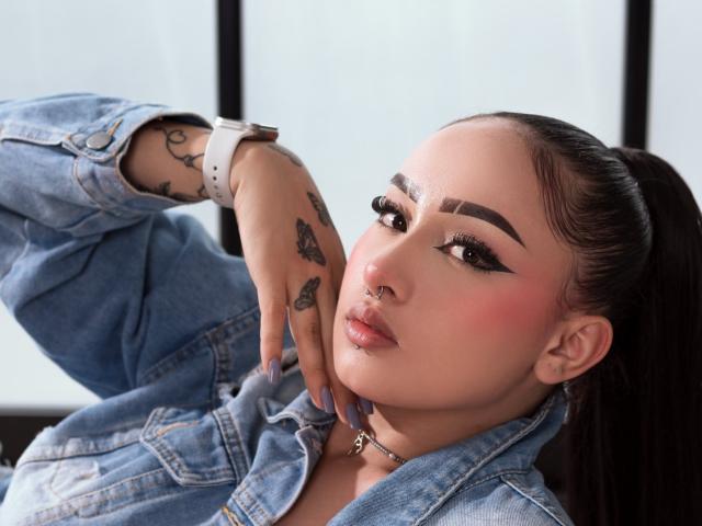 MelanieG with tattoos on her hand and wrist, wearing a blue denim jacket, white watch, and choker, has pink blushed cheeks, and is looking at the camera with a slight pout.