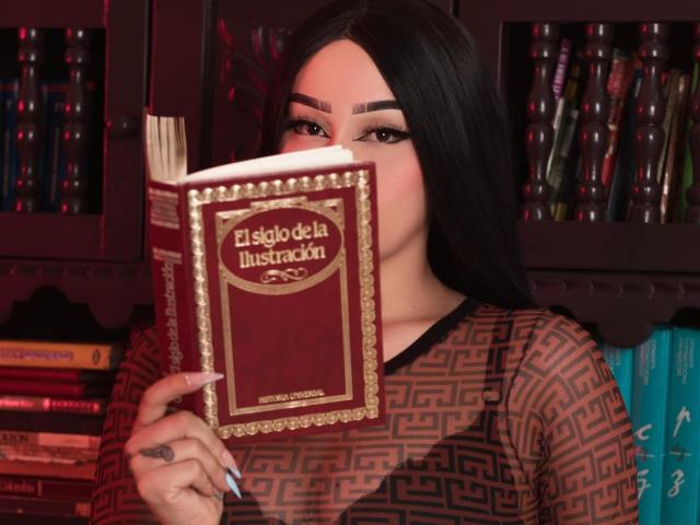 MelanieG with long black hair, holding a book in front of her face, wearing a patterned brown top, bookshelf in background, focused expression, nails painted blue.