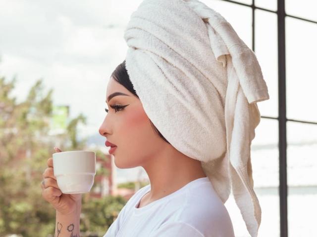 MelanieG with a white towel wrapped around her head, holding a white cup, wearing a white shirt, has dark hair and a tattoo on her right hand.