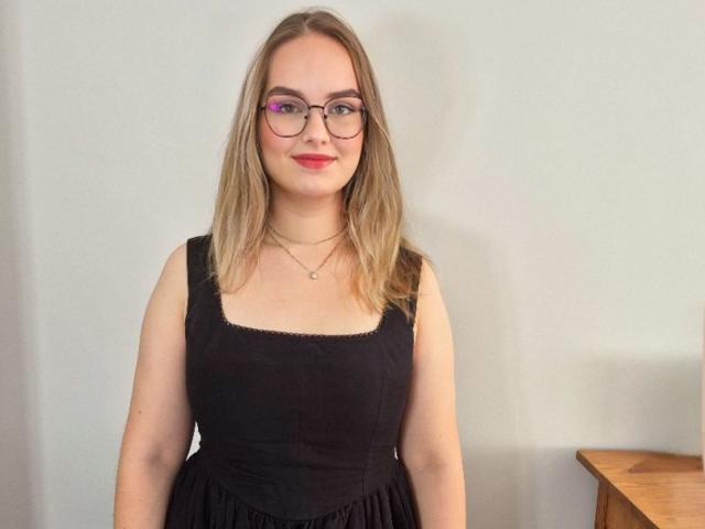 AliceNewX with medium tits, wearing a black dress, glasses, and necklace, smiling with light brown hair and red lipstick, standing against a plain wall.